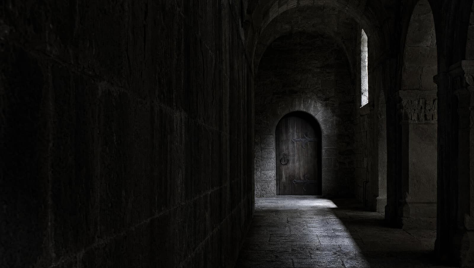 Dunkler Klostergang aus Stein, ein schmaler Lichtstreifen fällt auf den Boden vor einer eisenbeschlagenen Tür.